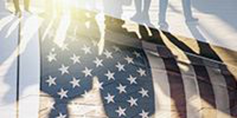 People standing in front of a light source, the silhouettes from their legs and feet are at the top of the image, while their shadows are casting onto a translucent American flag.
